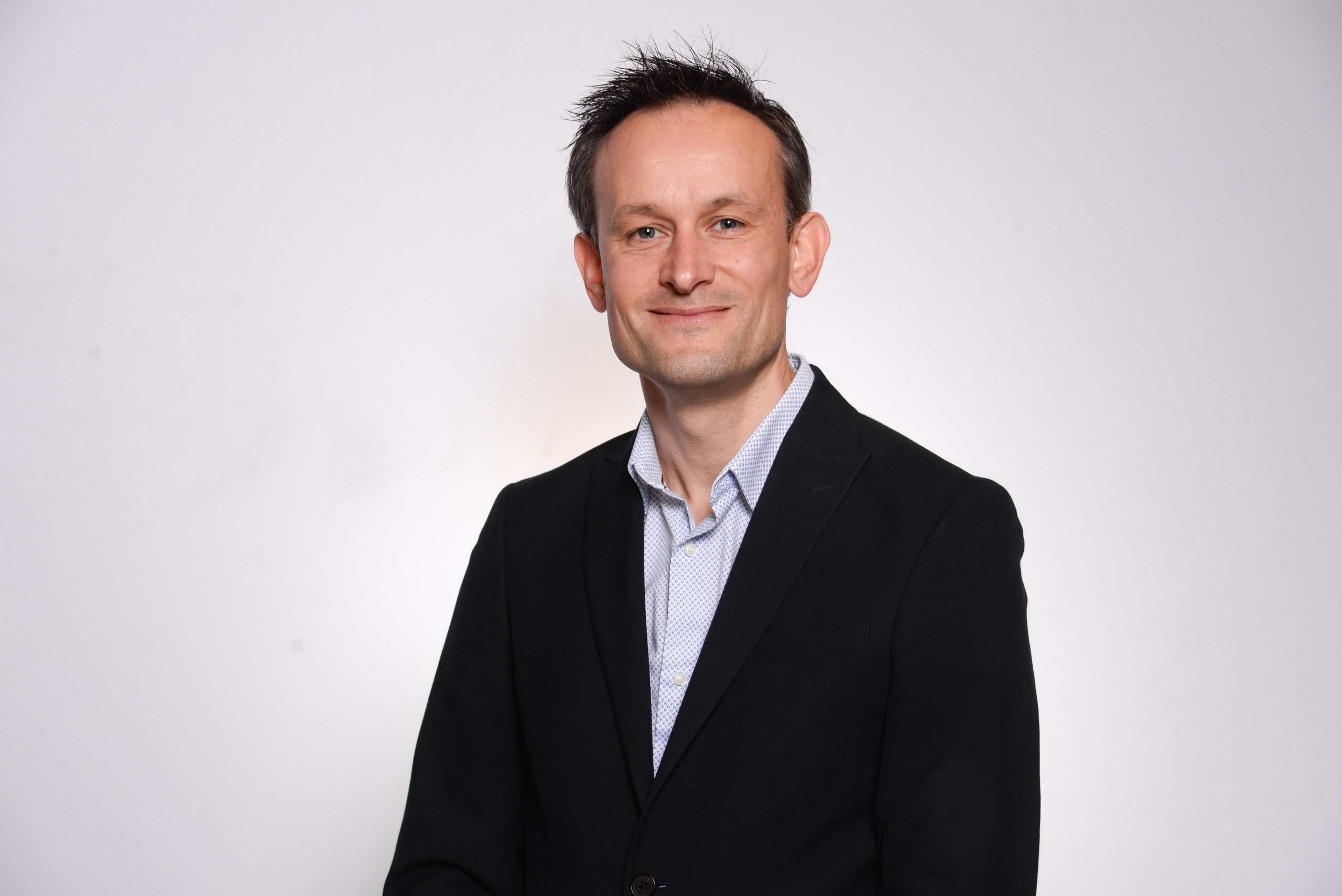 Professional businessman in a dark suit and light shirt posing on a plain white background, representing leadership, executive, corporate, and professional consulting services.