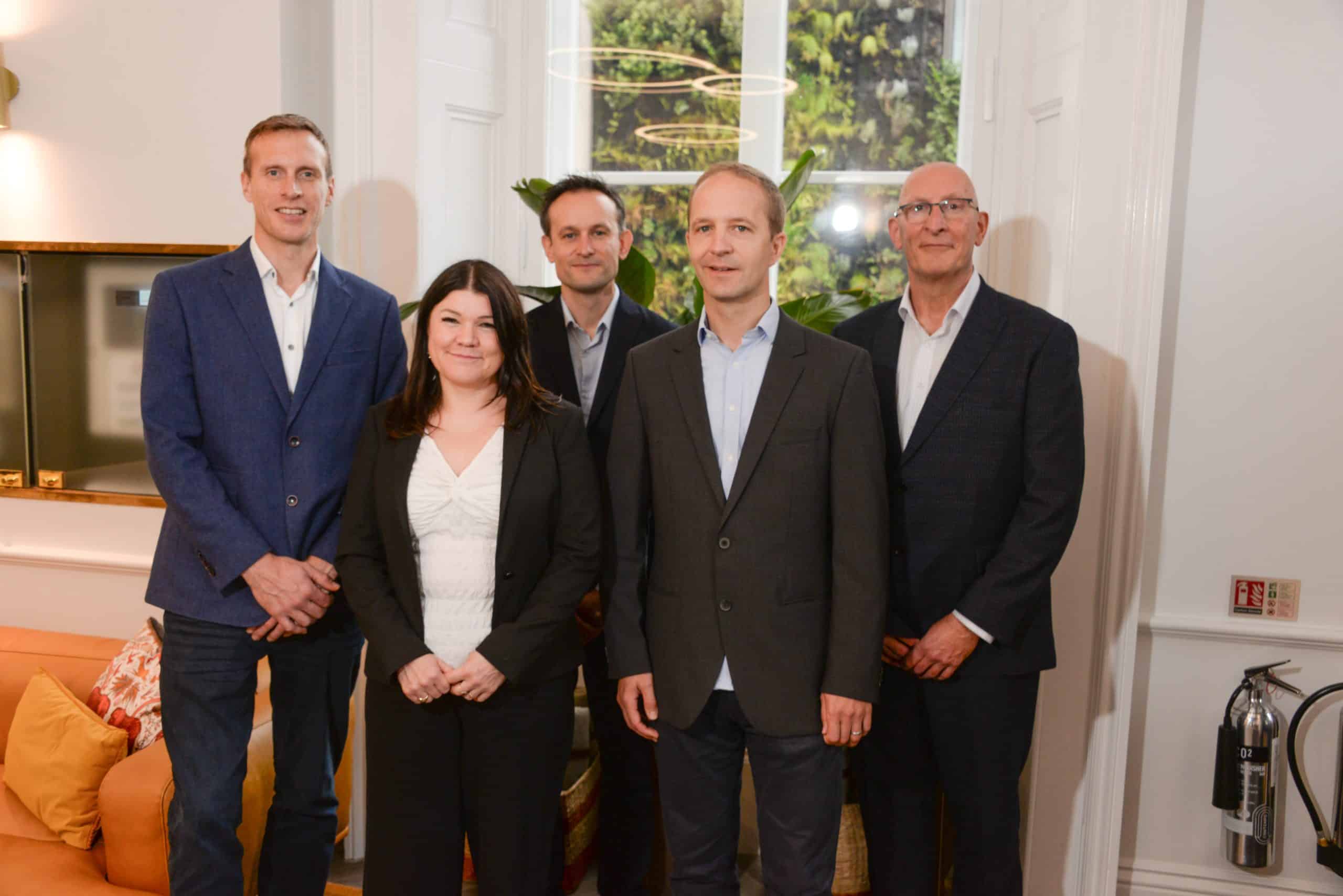 Professional team photo of business professionals standing indoors at an enterprise event or conference, with modern decor and greenery visible through the large window behind them.